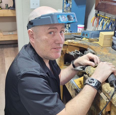 Technician wearing a headlamp works at a cluttered jewelry bench, handling tools and materials.