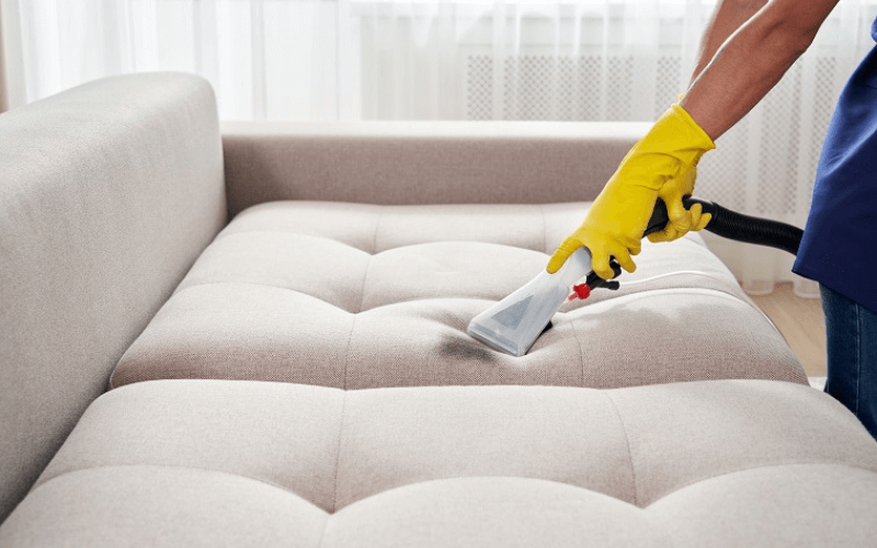Person in yellow gloves cleaning a beige tufted couch with a handheld upholstery cleaner.