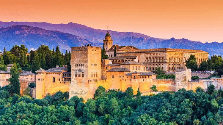 Het paleiscomplex van het Alhambra bij zonsondergang, met de gouden muren, torens, bomen en bergen op de achtergrond.
