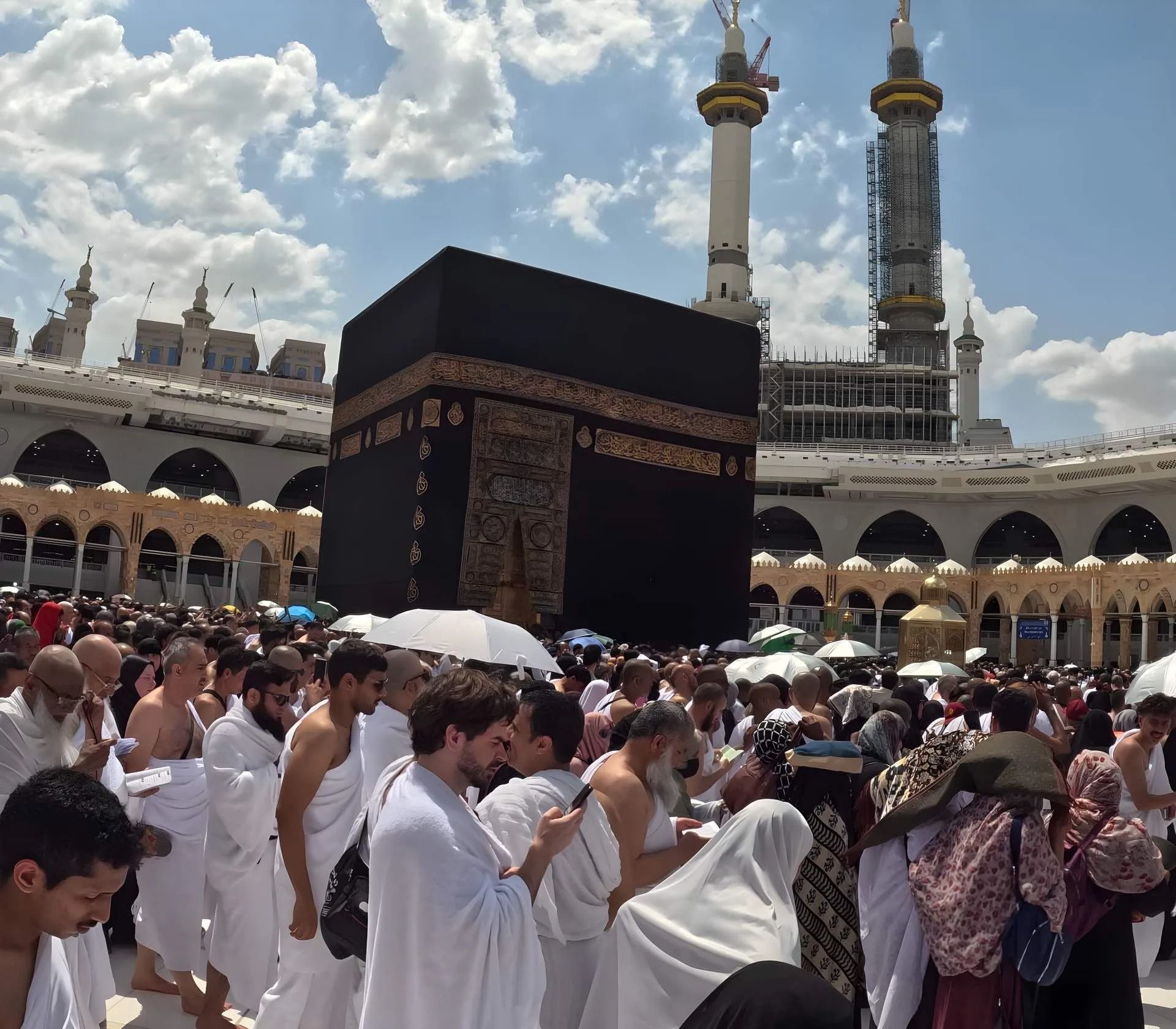 Pelgrims in witte gewaden cirkelen rond de Kaaba in de Grote Moskee in Mekka, met minaretten en een bewolkte hemel.