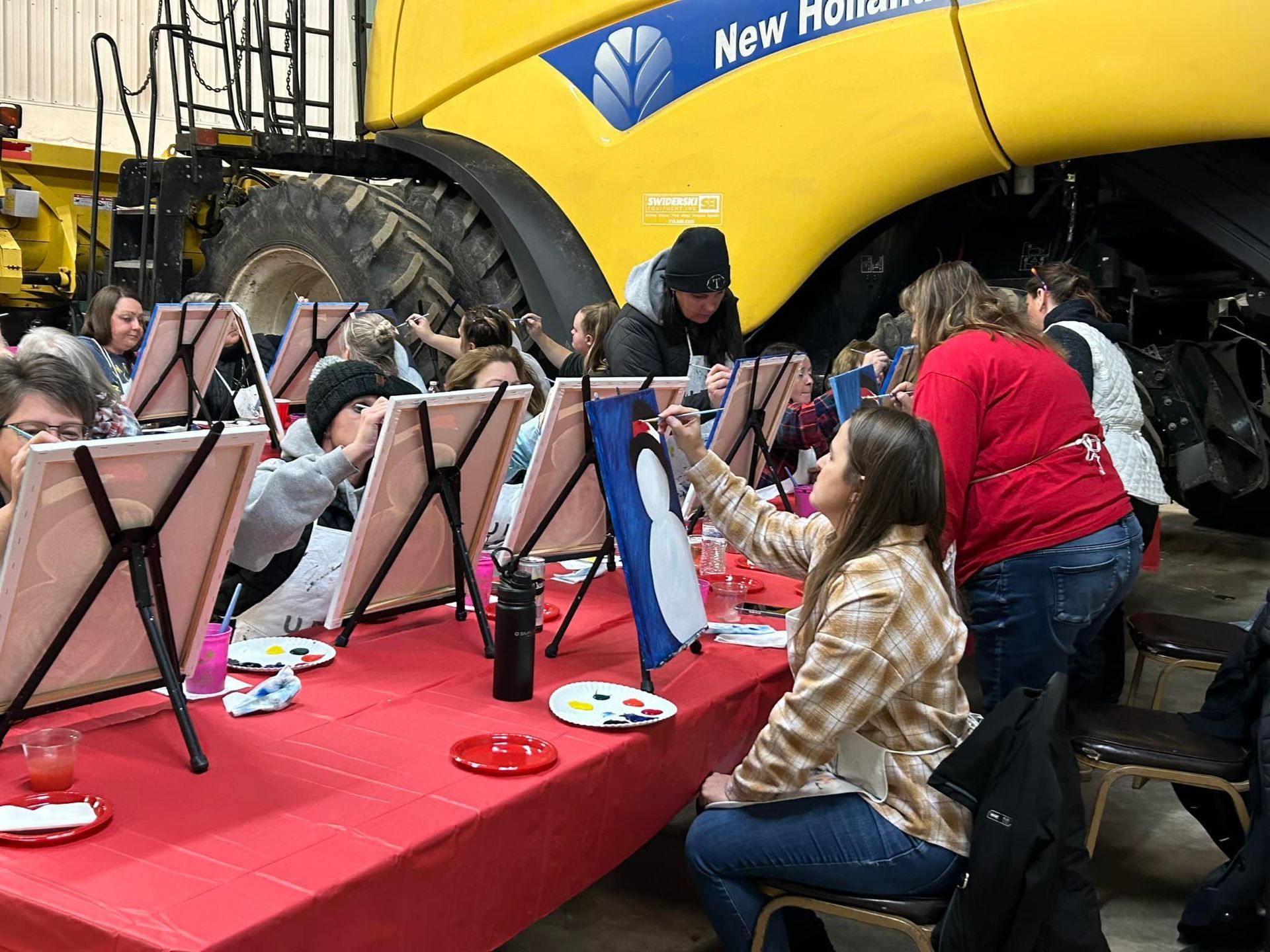 A group of people are sitting at a table painting in front of a yellow tractor.
