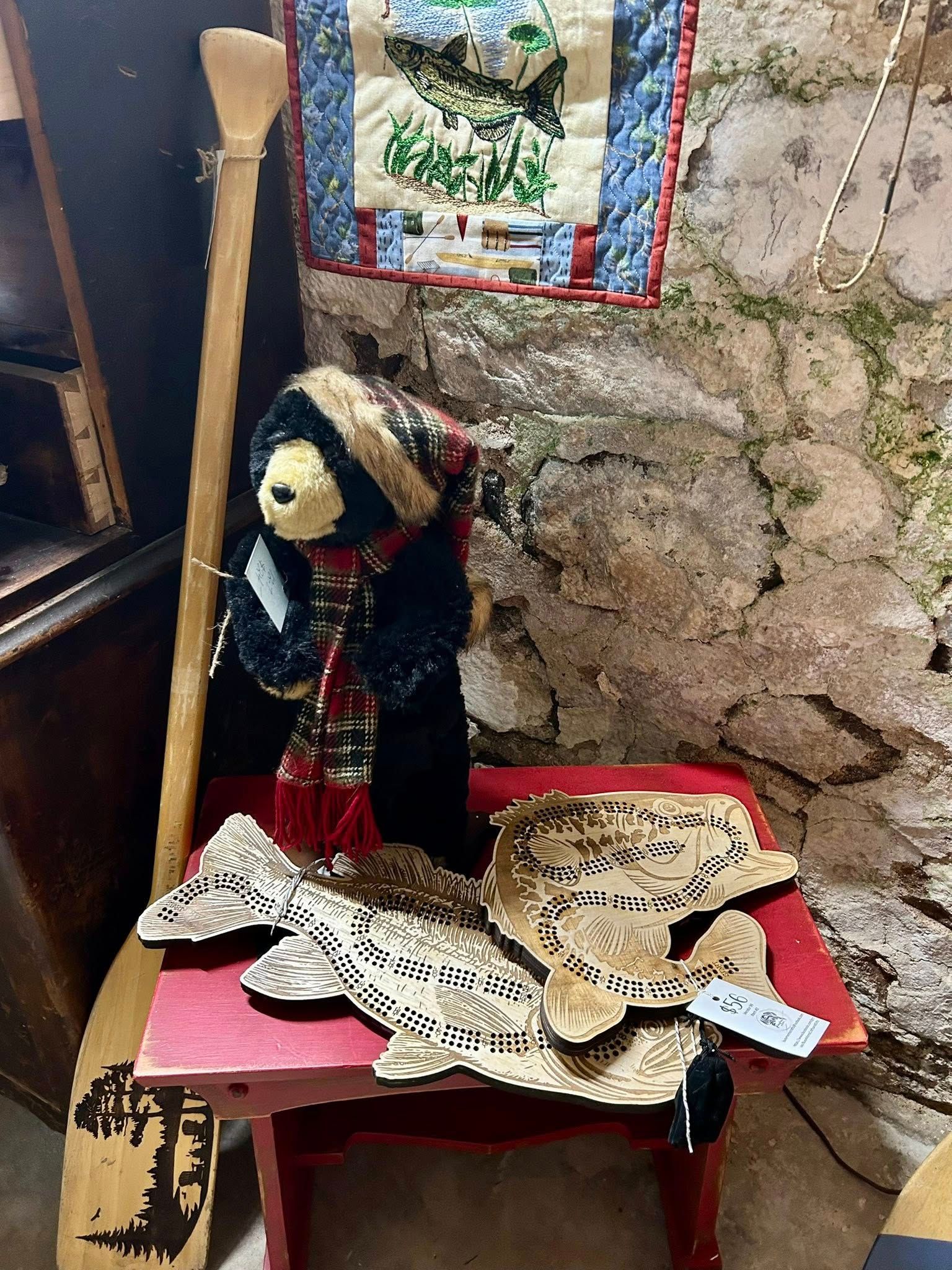 Bear in scarf, wooden fish and paddle on a red table, against a stone wall. Quilt above.