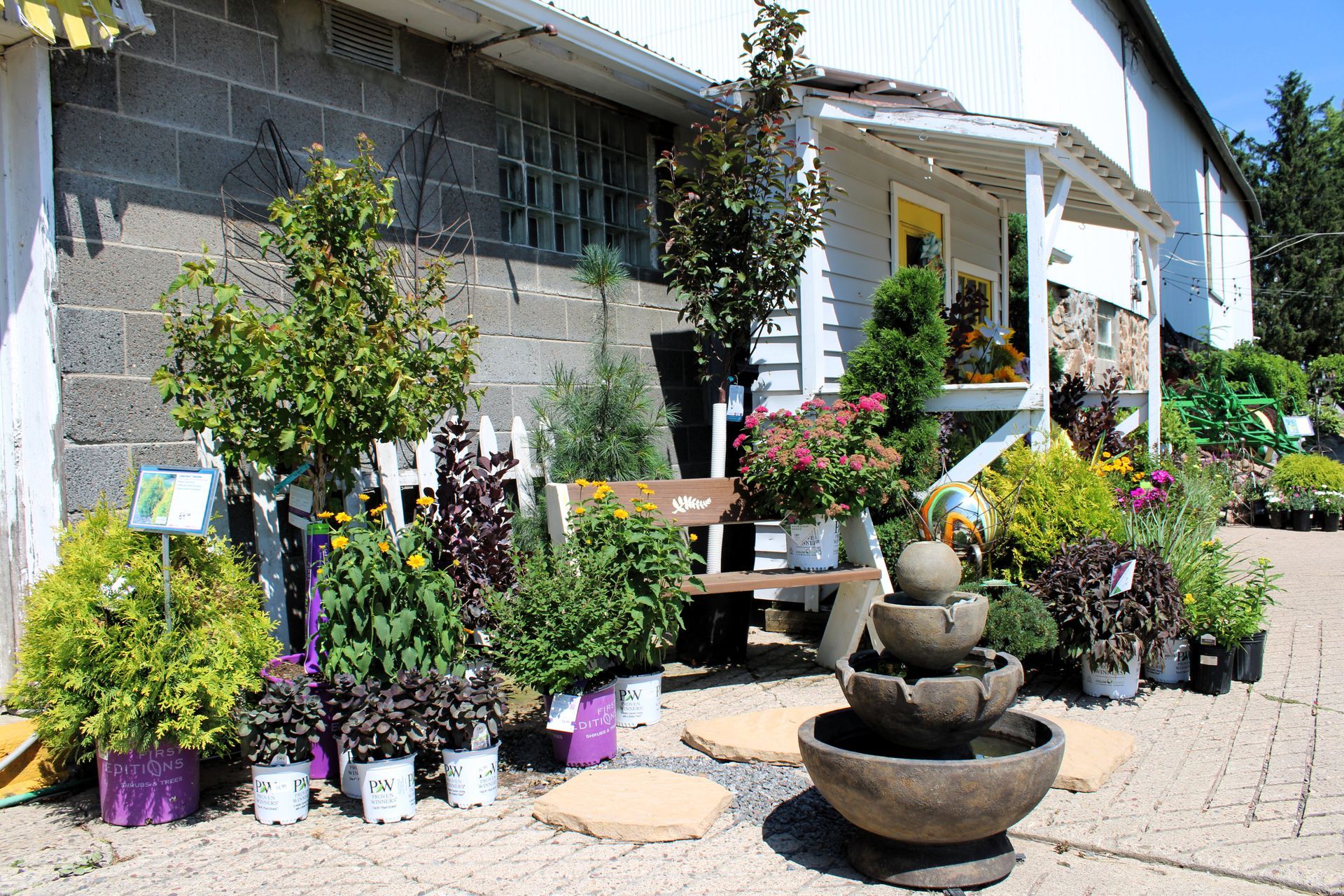 A garden center entrance with plants in pots, a small fountain, and a bench on a paved area.