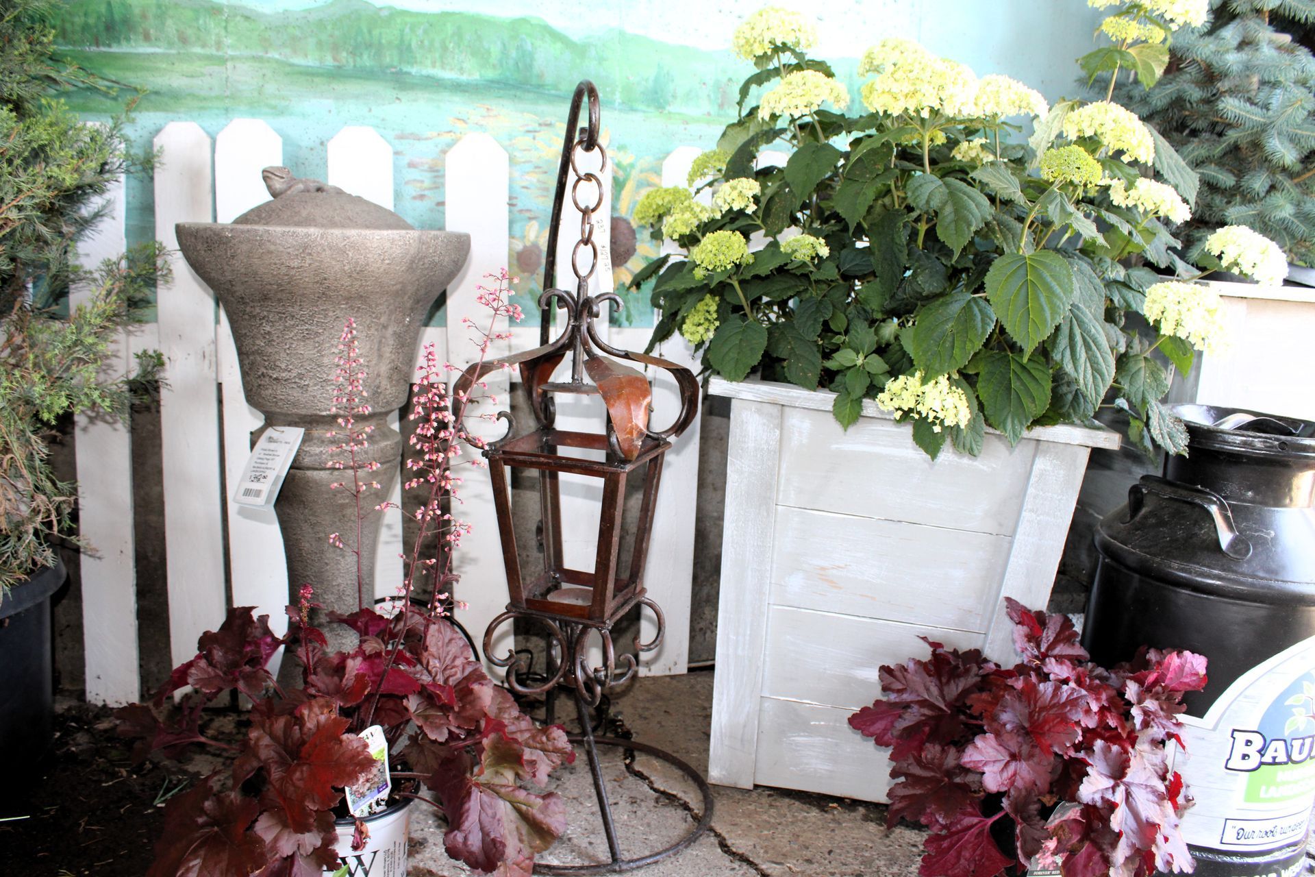 Outdoor display with decorative items: lantern, planter, and red foliage in front of a white picket fence and plants.