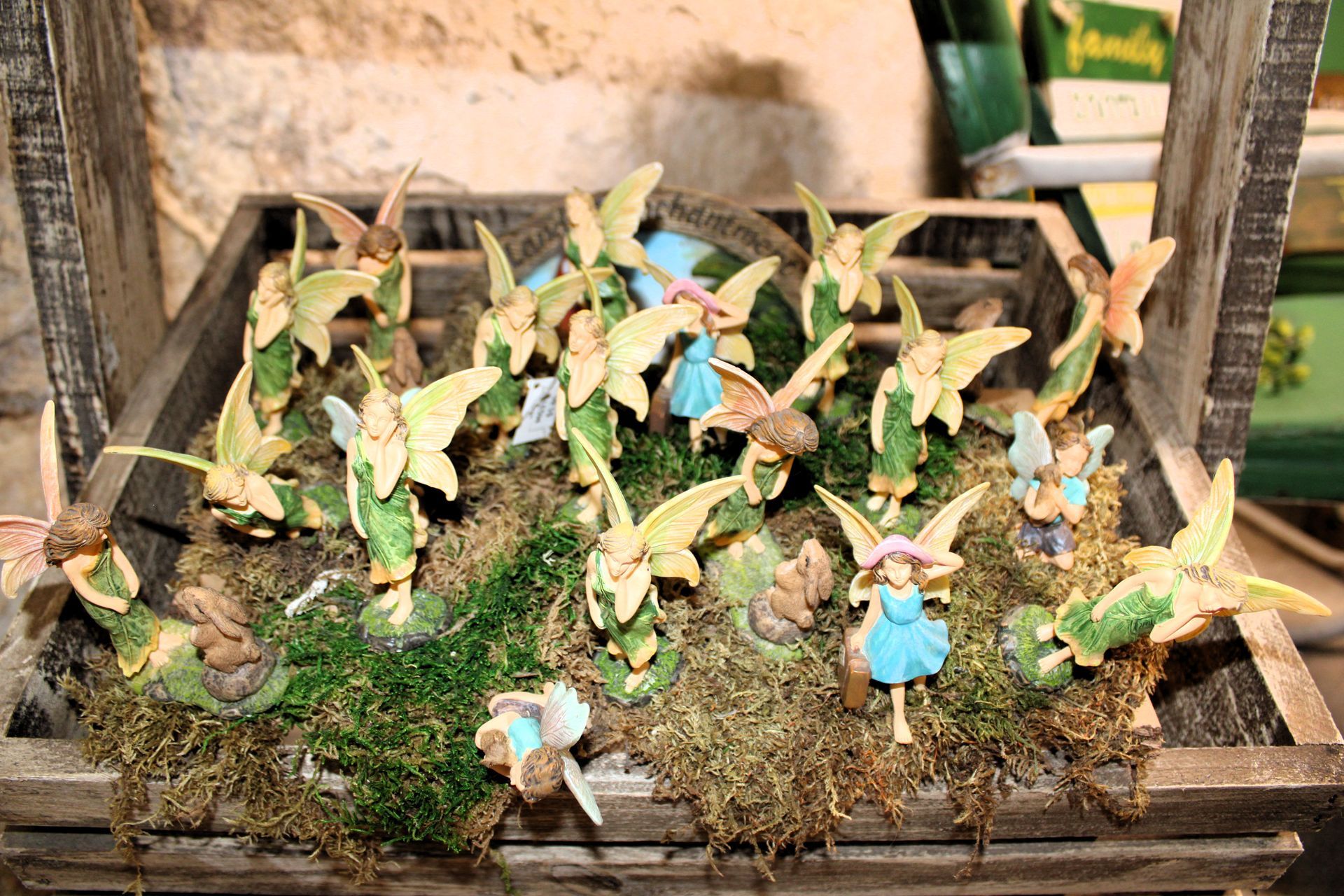 Wooden crate filled with fairy figurines, moss, and greenery. Light yellow wings, blue and pink outfits.