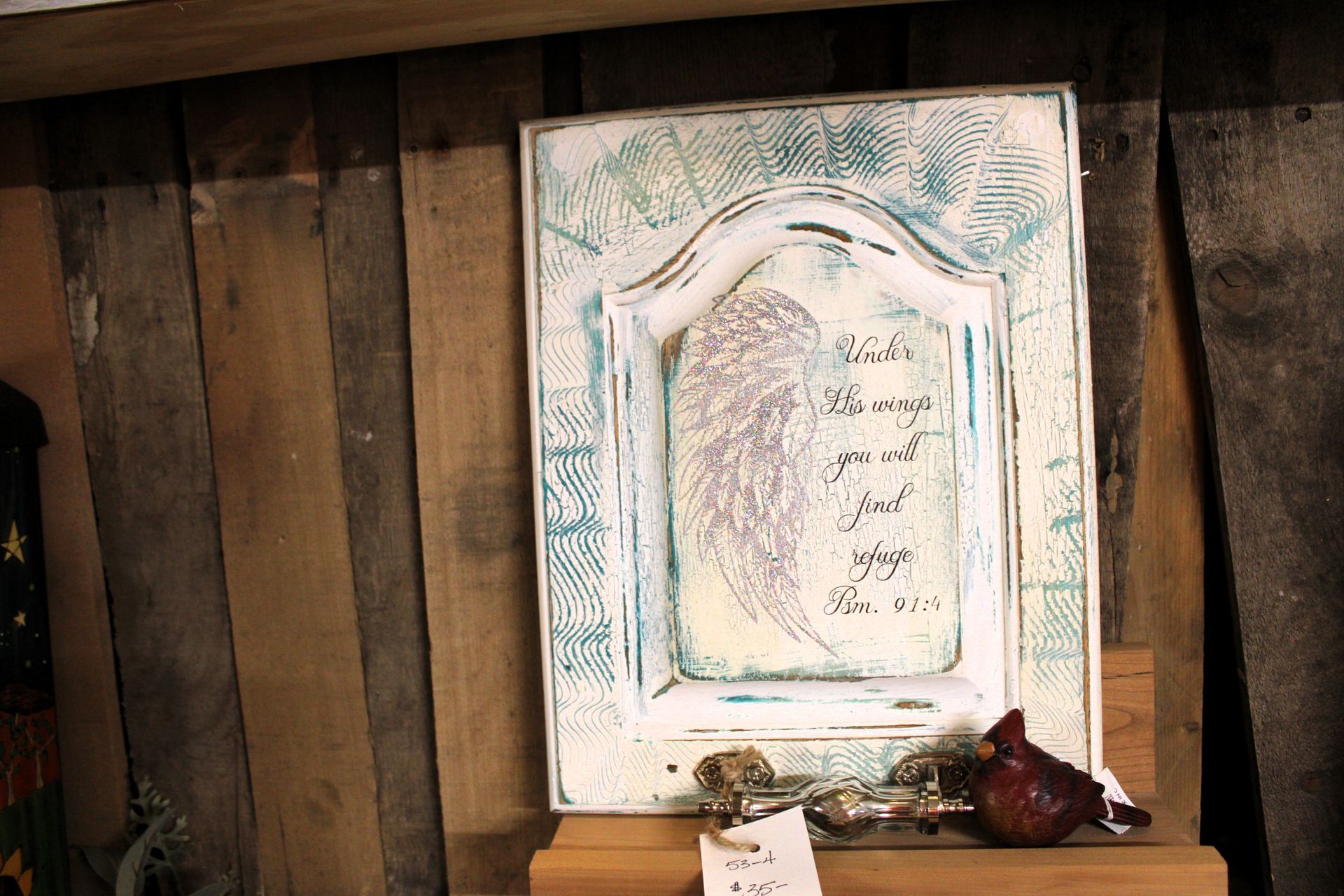 White painted wooden wall art with a wing design and text, resting on a wooden shelf in front of a rustic wooden wall.