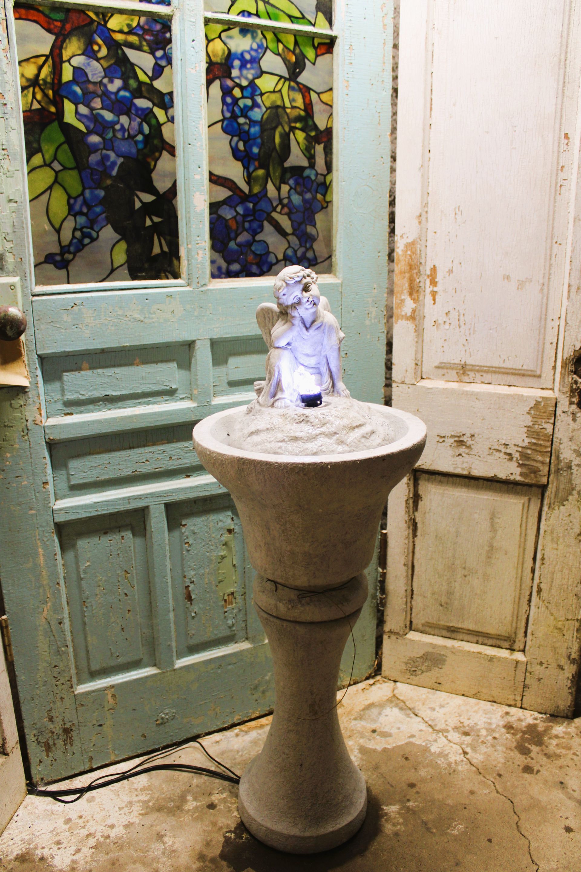 Stone fountain with angel statue, lit up, near a weathered blue door with stained glass.