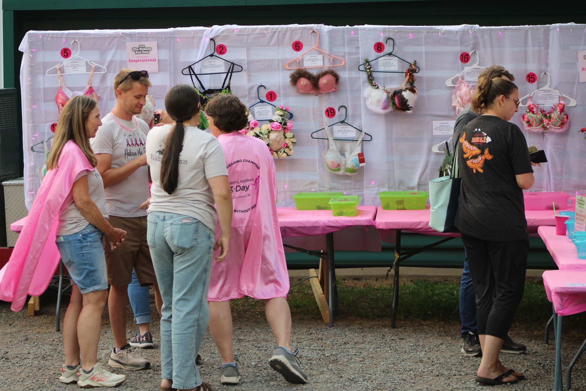 People looking at decorated bras displayed on a wall, with pink tables and capes, outdoors.