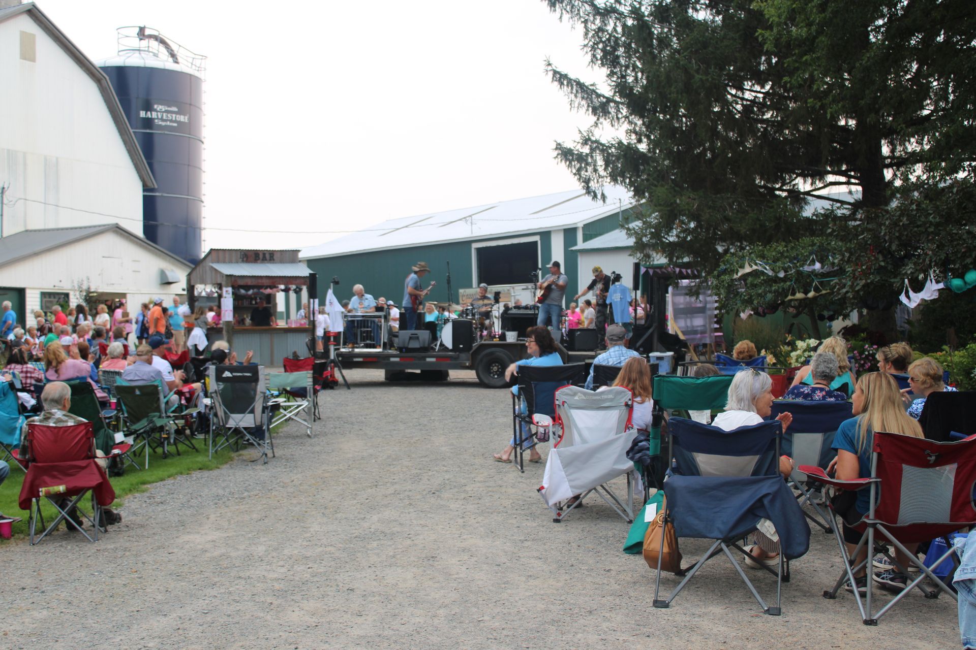 Outdoor concert at a farm: band on a trailer stage, audience in folding chairs, white buildings, silo in background.