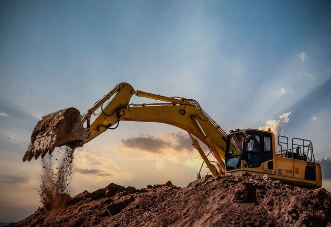 Yellow excavator digging a dirt pile at sunset under a dramatic sky