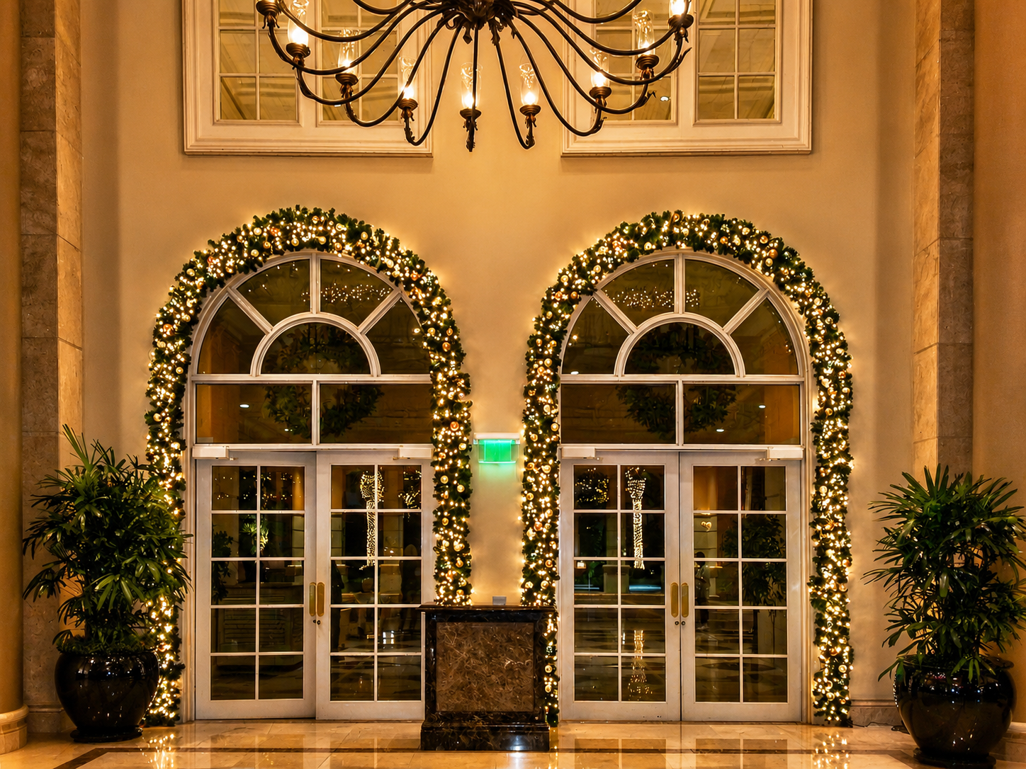 Illuminated hotel entrance with twin arched glass doors, holiday garland, and chandeliers above.