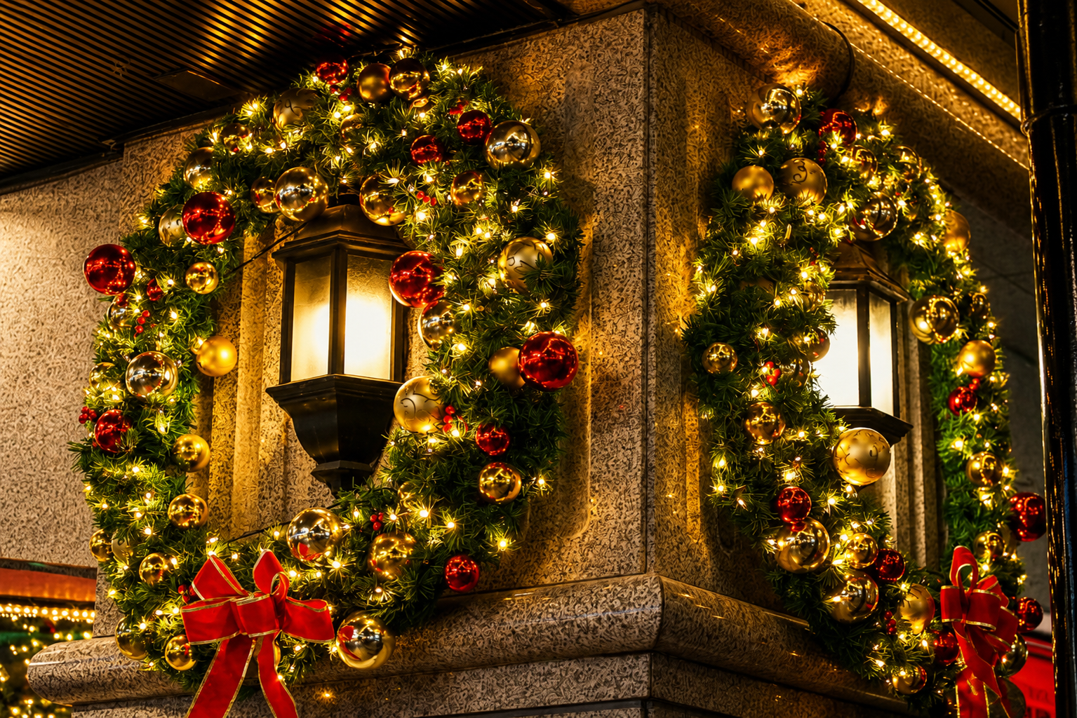 Christmas wreaths with red bows and warm lights around corner lanterns on a decorated building exterior
