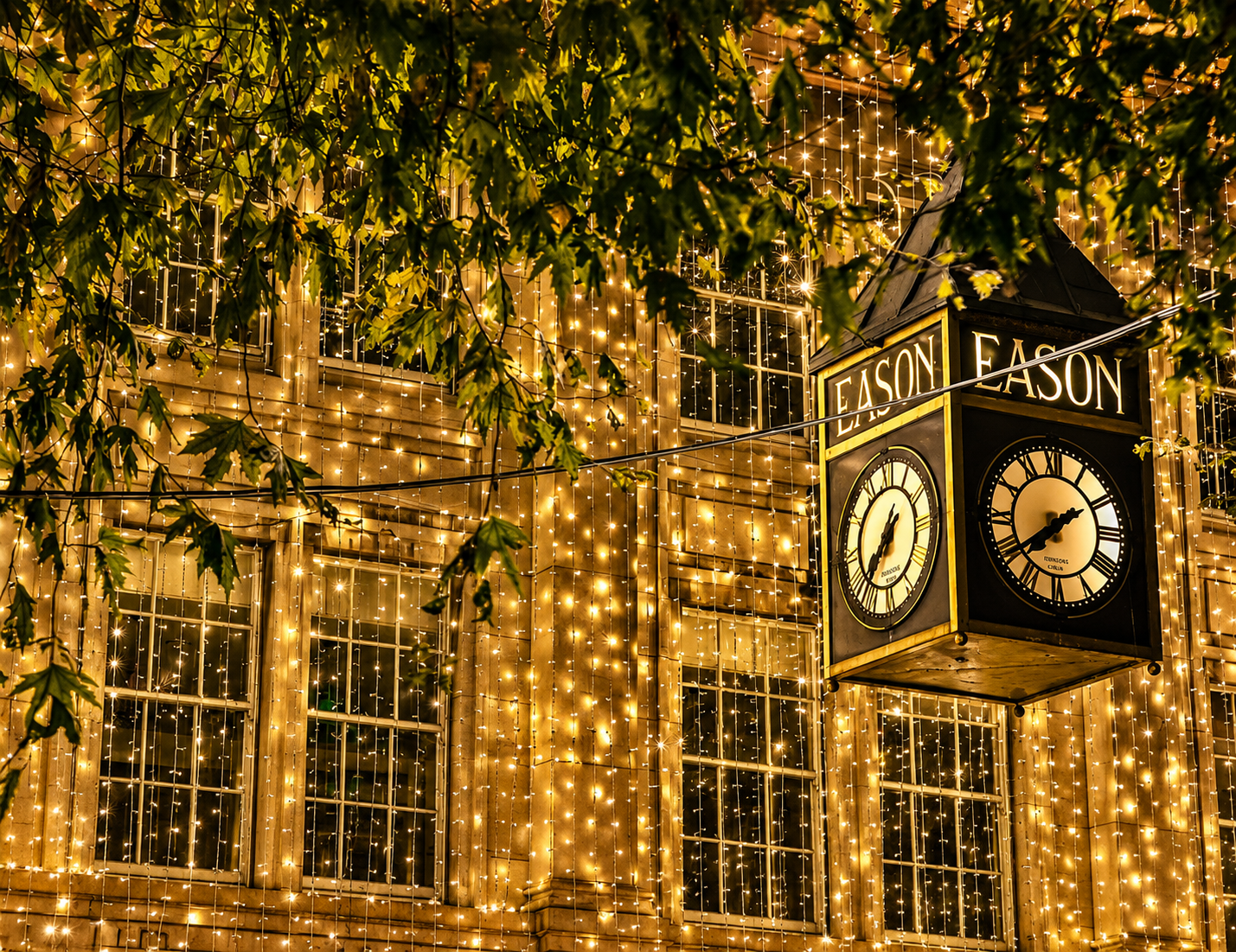 Clock sign hanging above a building draped in golden holiday lights at night