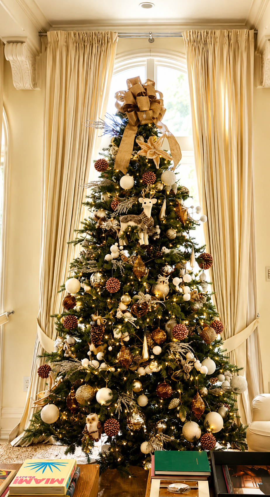 Tall decorated Christmas tree with gold ornaments and lights in a bright room by curtained windows