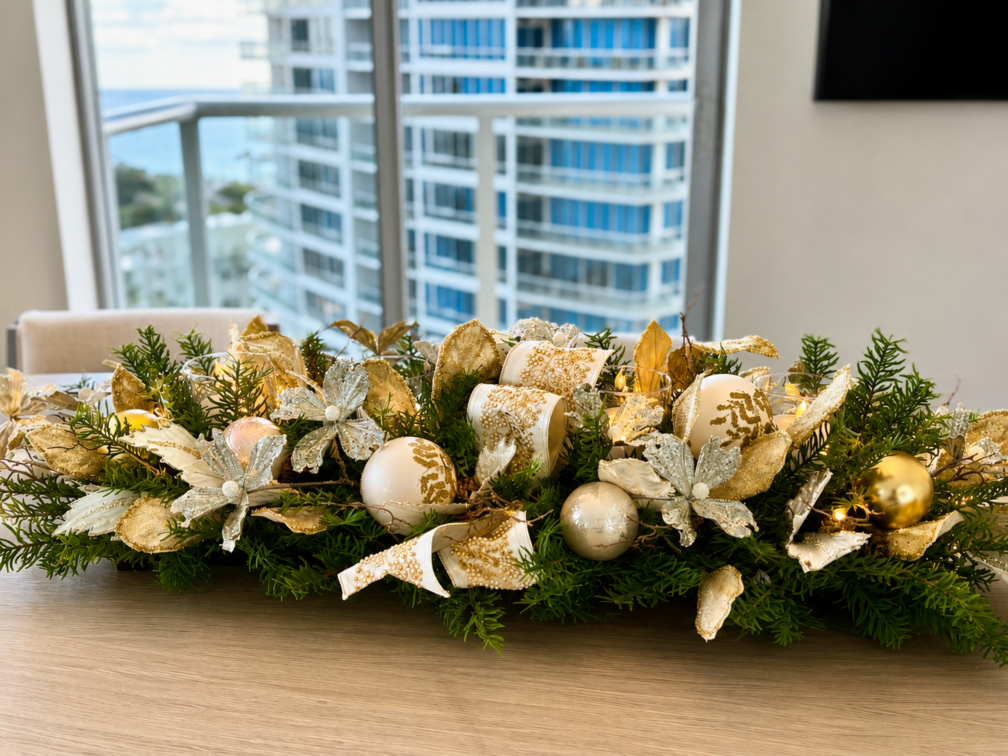 White and gold holiday centerpiece with pine, ornaments, and candles on a table before a balcony view