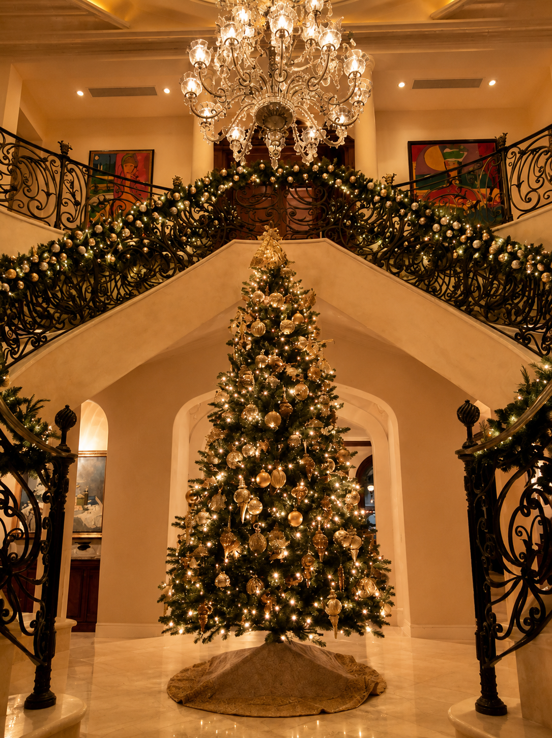 Grand staircase decorated with garlands and chandelier above a lit Christmas tree in a grand foyer