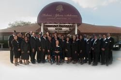 Group of people in black formal attire posed outside a building with a maroon awning.