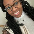 Woman with locs, glasses, smiling, wearing turtleneck and patterned shirt.
