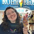 Person smiling, holding an ice cream cone with sprinkles in front of Kohr Bros Frozen Custard shop.