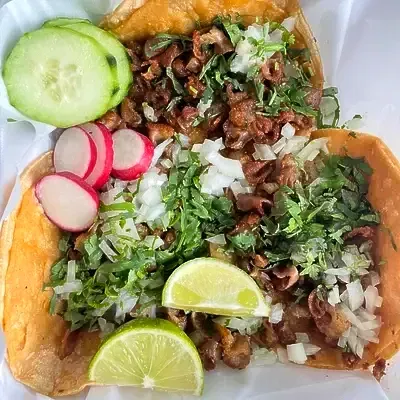 Two tacos topped with meat, cilantro, onions, lime, radishes, and cucumber.
