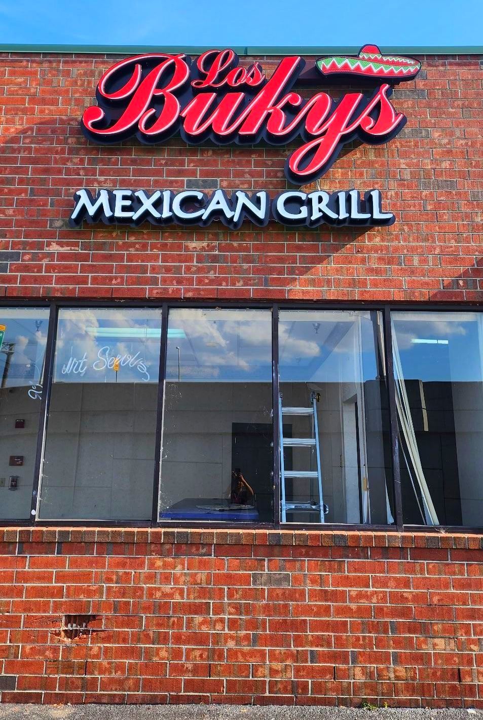 Exterior of Los Baky's Mexican Grill restaurant with red brick facade, red and white sign, and a ladder inside.