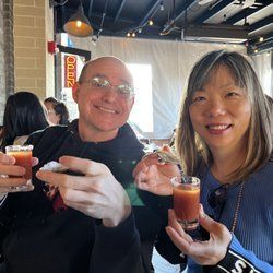 Two people smiling, holding glasses of red liquid indoors, possibly a restaurant.