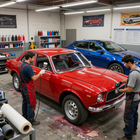Taller de carrocería con un coche clásico rojo en reparación, un coche azul al fondo y dos mecánicos cerca.