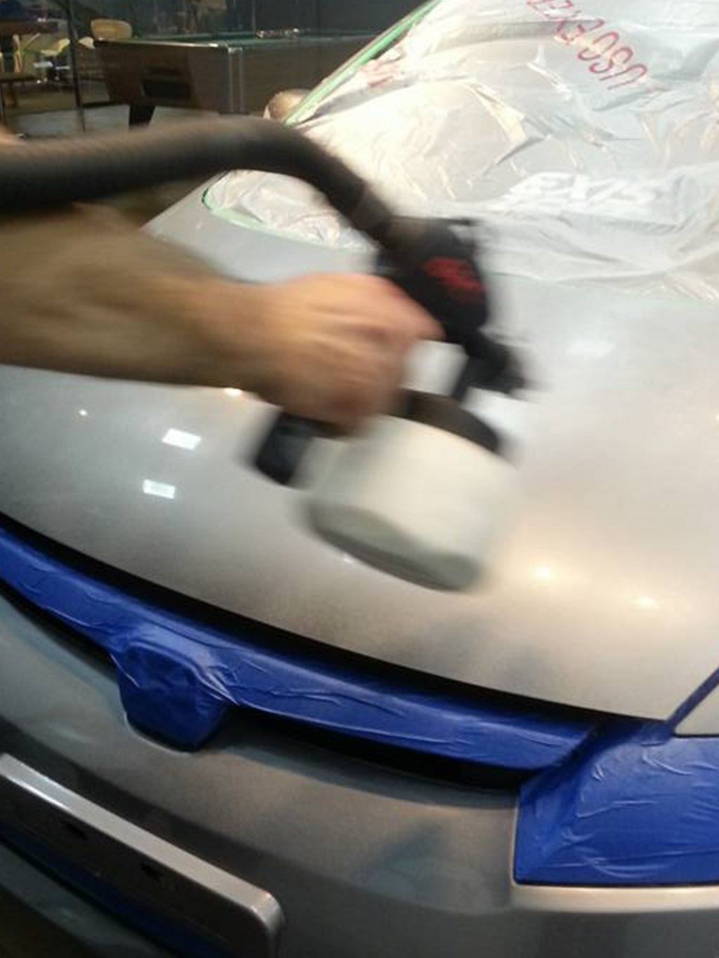 Una persona pule el capó plateado de un coche con una pulidora; la zona de la parrilla delantera está cubierta con cinta adhesiva azul.