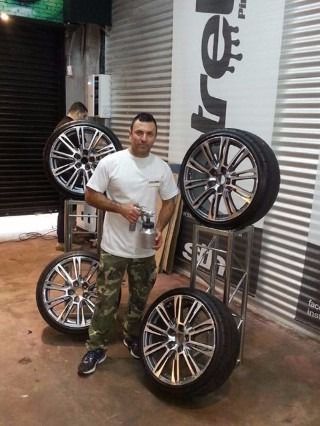 Hombre de pie junto a una pila de ruedas de coche en una tienda, sosteniendo una bebida, con un gran letrero vertical al fondo.