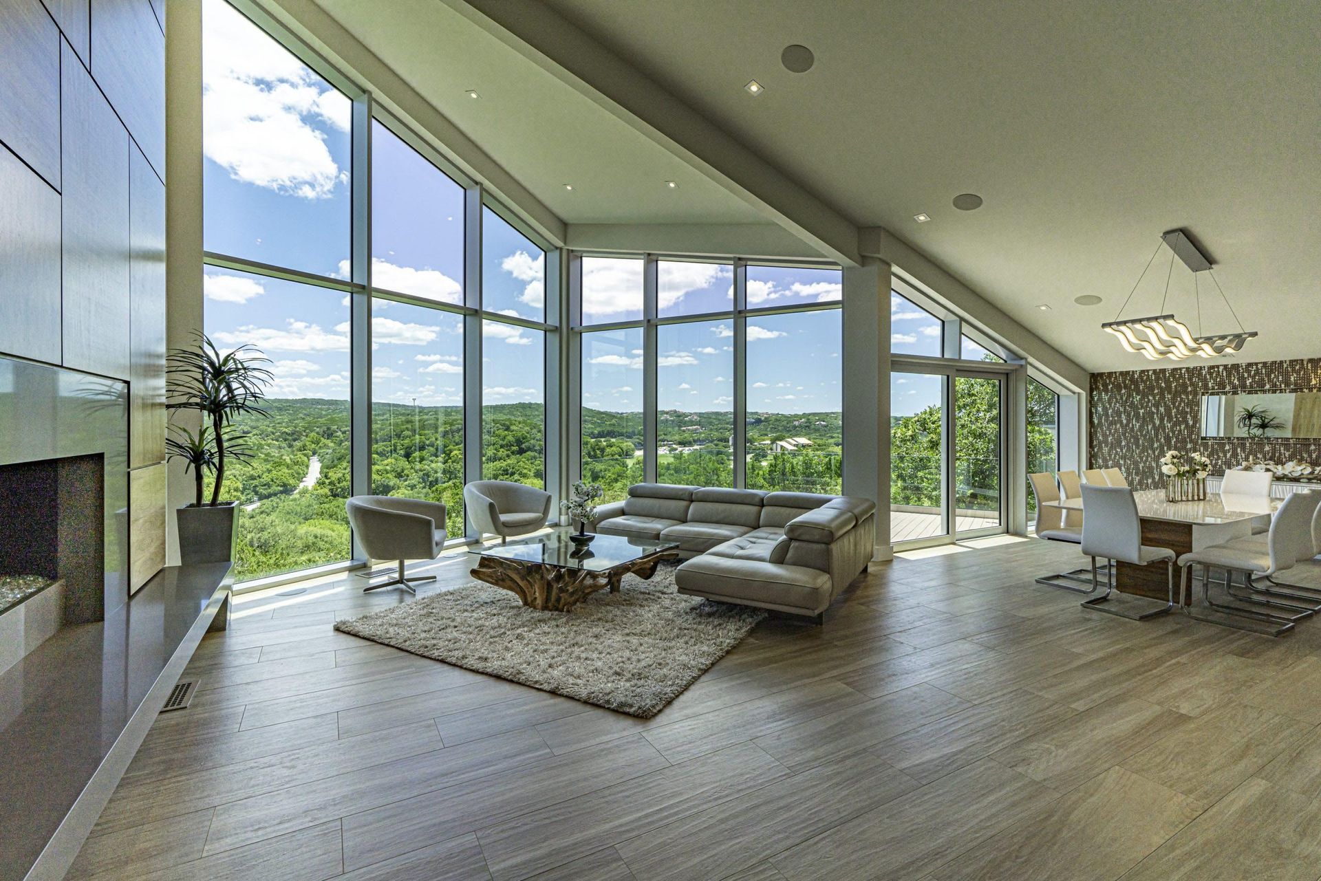 Modern living room with floor-to-ceiling windows, overlooking a green landscape. Features a fireplace, seating area
