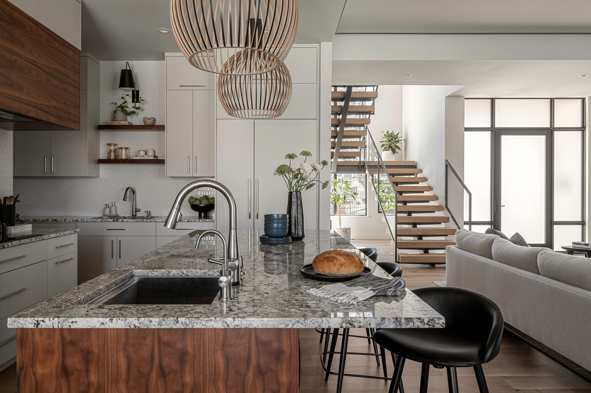 A modern kitchen with a granite island, walnut cabinetry, wooden pendant lights, and a staircase in the background.