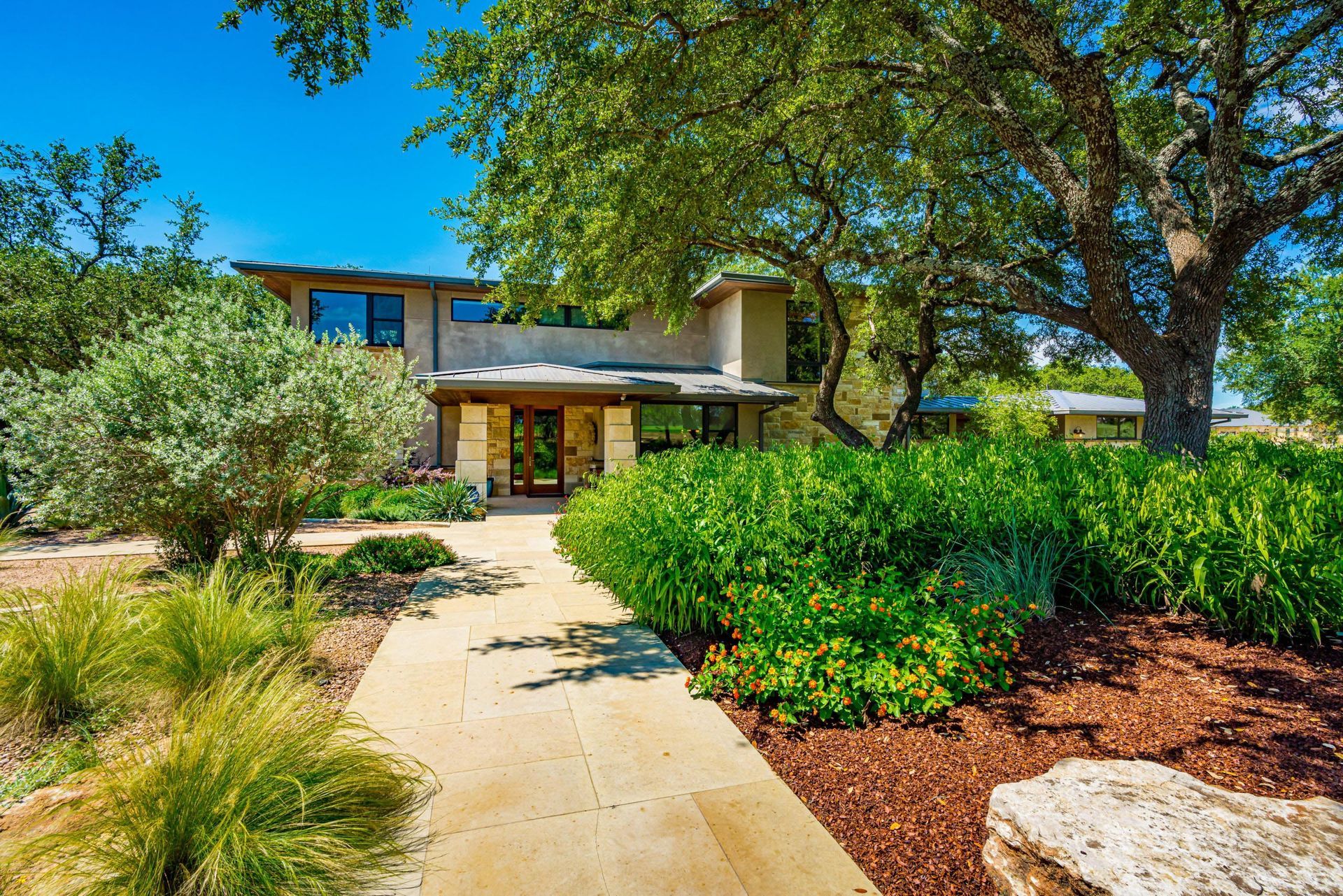 Stone path leads to a house with a wooden door, surrounded by lush green shrubs and trees, under a blue sky.