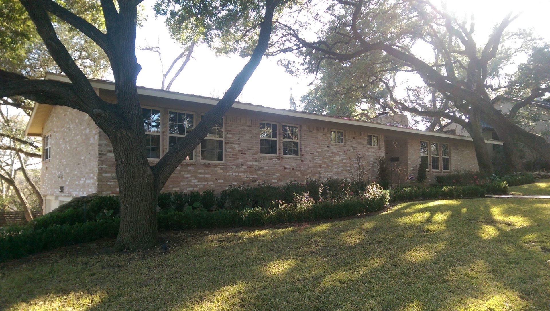 Ranch-style house with brick and stone facade, nestled under large trees on a grassy hill.