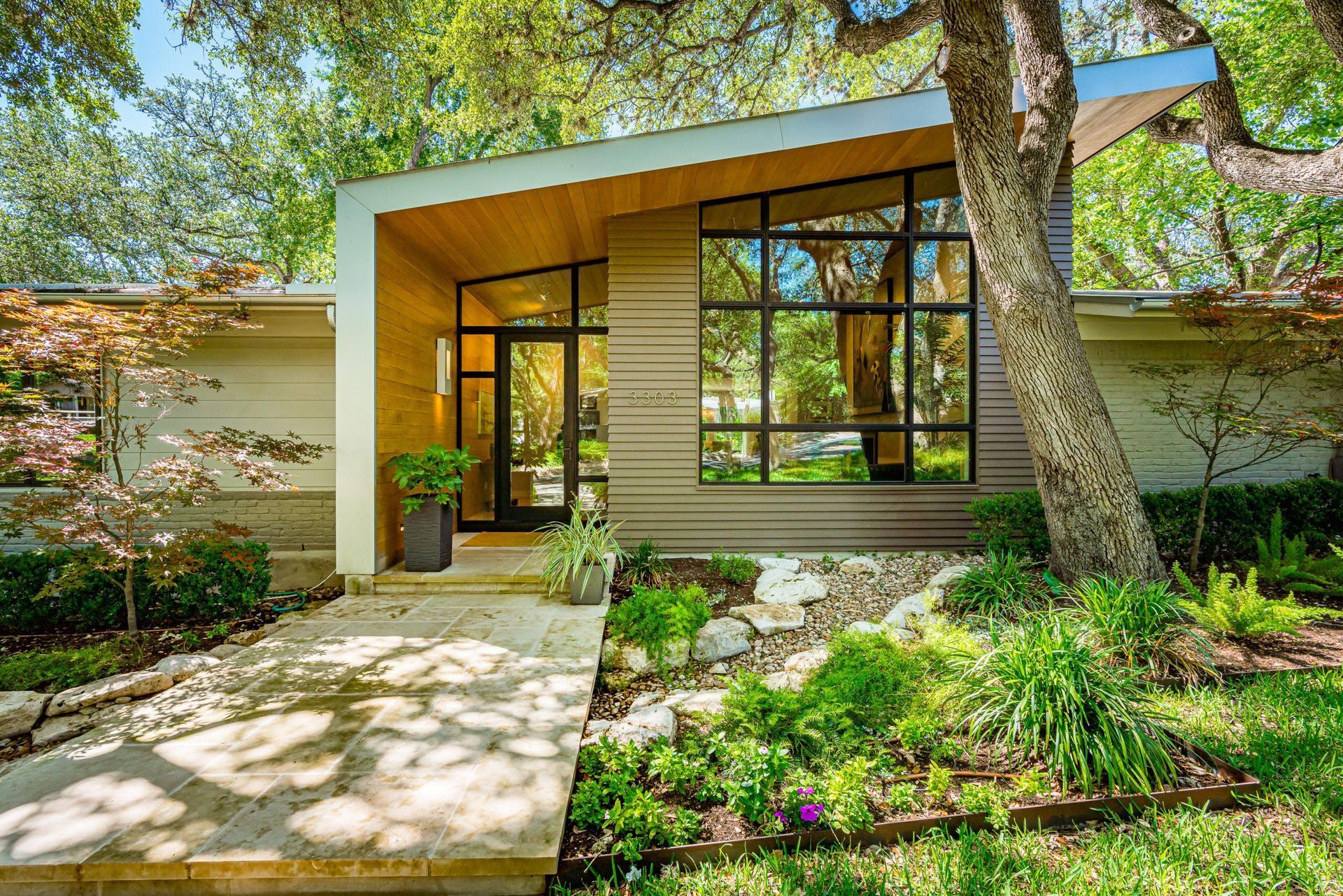 Modern mid-century style home with a flat roof, large glass windows, and a stone walkway surrounded by lush trees.