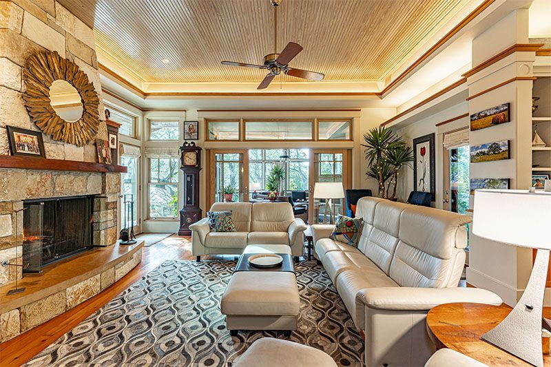A bright living room with cream leather sofas, a stone fireplace, a patterned area rug, and a ceiling fan.