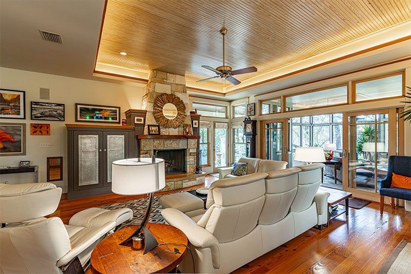 A spacious, warm-toned living room featuring white leather sofas, a stone fireplace, wood ceiling, and large glass doors.