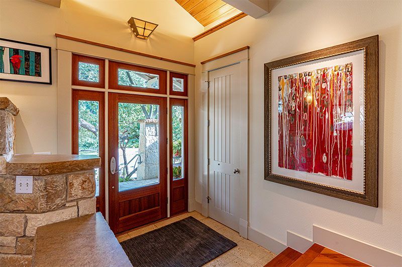 A bright entryway featuring a wood-framed front door with sidelights, a stone pillar, a white door, and abstract red art.
