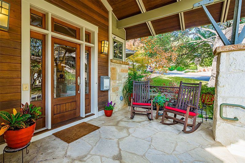 A covered stone porch featuring two wooden rocking chairs with red cushions, a potted plant, and a wooden front door.
