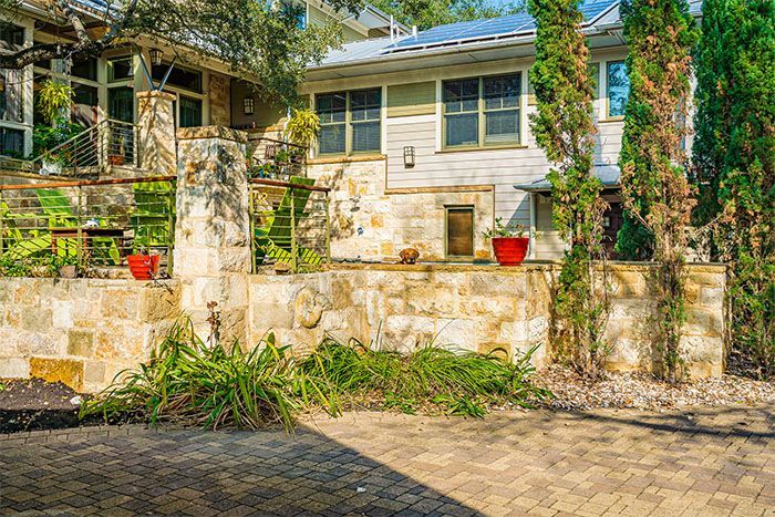 A stone patio and retaining wall in front of a light-colored house with tall cypress trees and potted plants.
