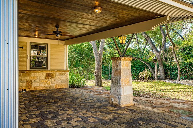 A covered outdoor patio with a stone column, wood ceiling, ceiling fan, and multi-colored paver floor looking onto trees.