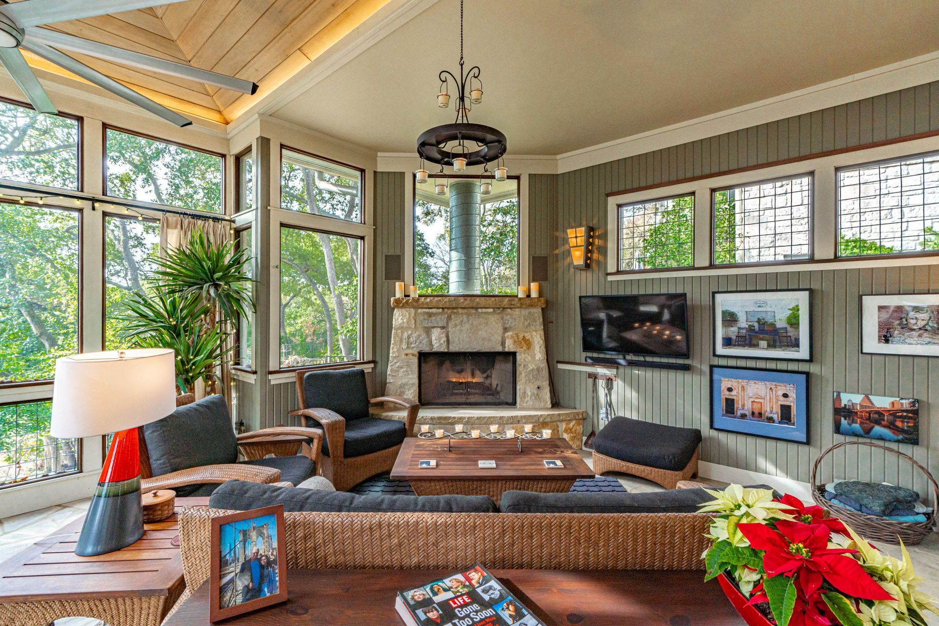 Cozy sunroom with stone fireplace, windows overlooking trees, wicker furniture, and a television.