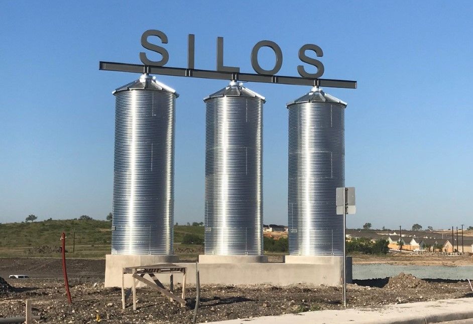 Three tall, silver corrugated metal grain silos stand in a row under a clear sky, topped with a sign reading