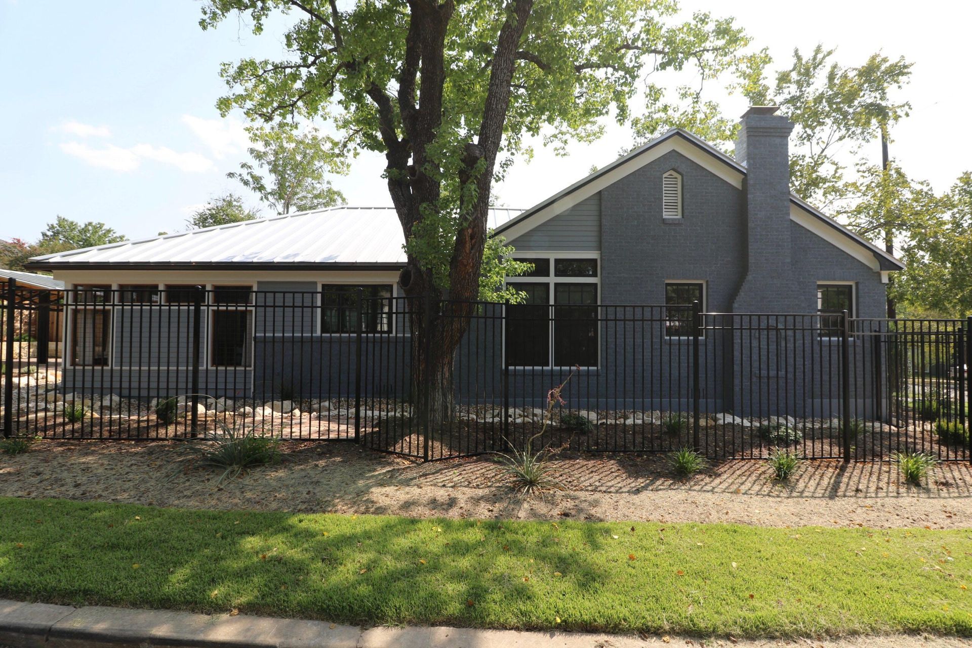 Blue house with black fence, metal roof, and large tree in front.