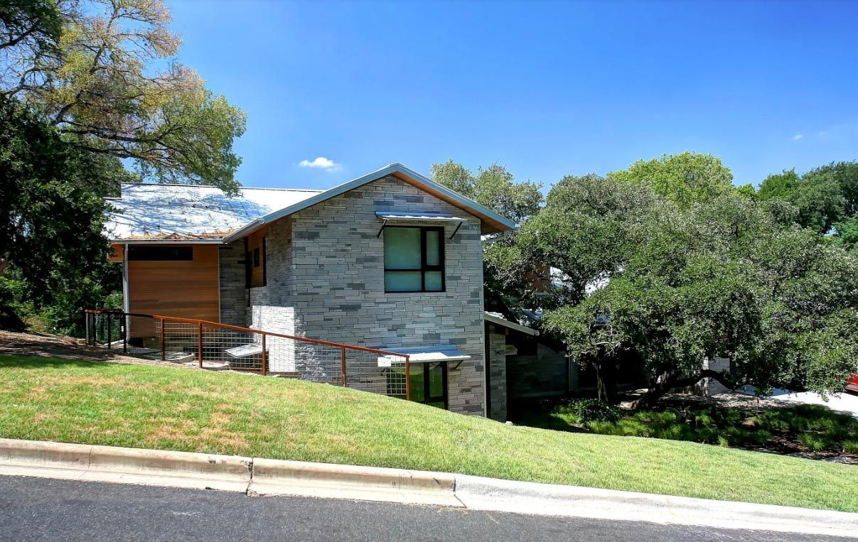 Stone and wood house on a grassy hill; blue sky; mature trees.