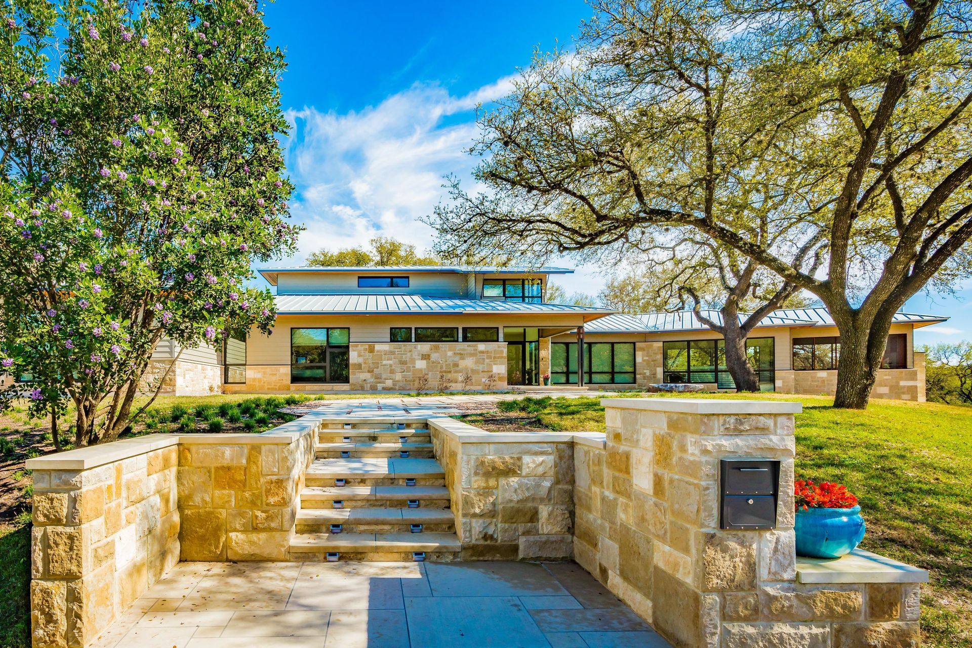 Modern home with stone facade, steps leading up, blue sky, and large tree.