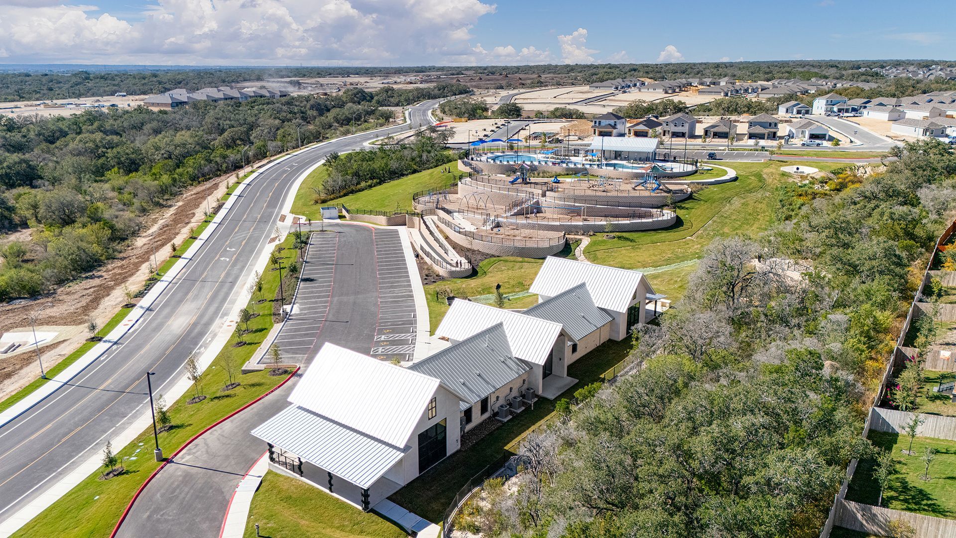 An aerial view of a white, multi-gabled building with a parking lot next to a curving road in a suburban, wooded area.