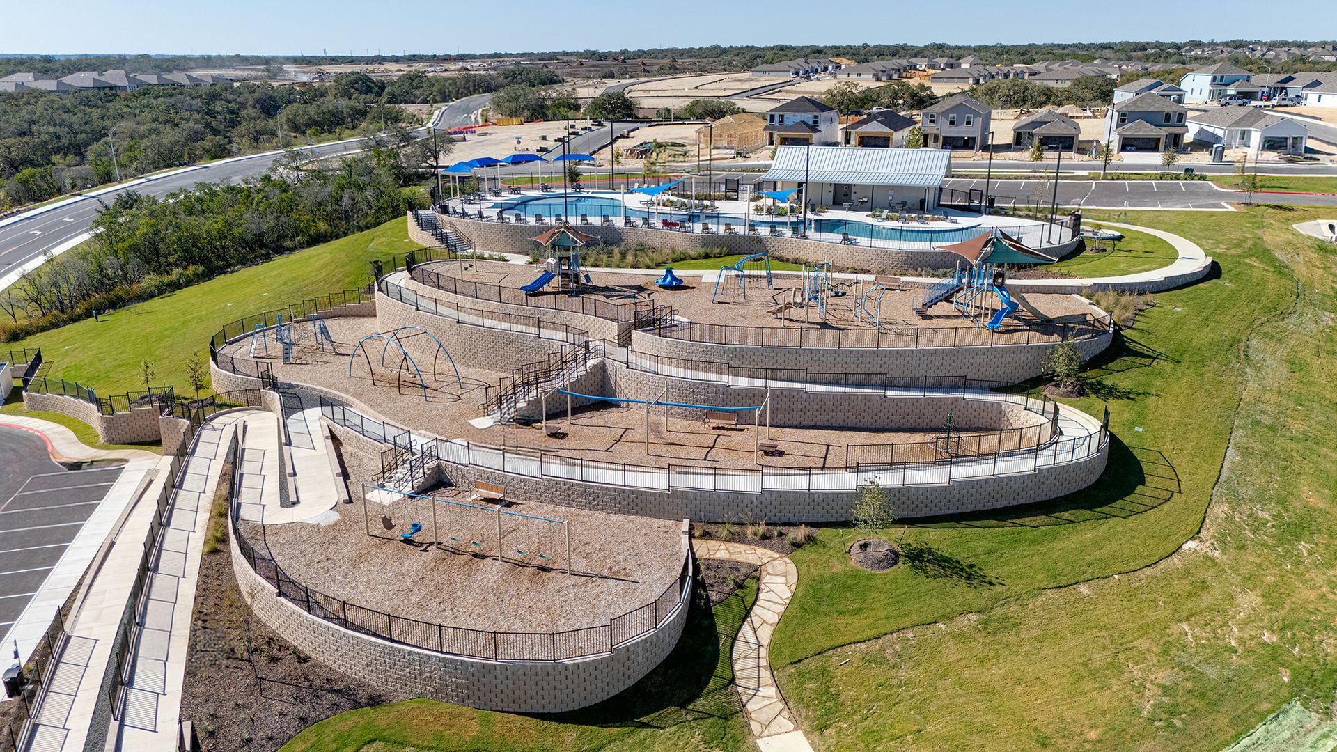 Aerial view of a terraced playground with slides and climbing structures, situated next to a community pool and parking.
