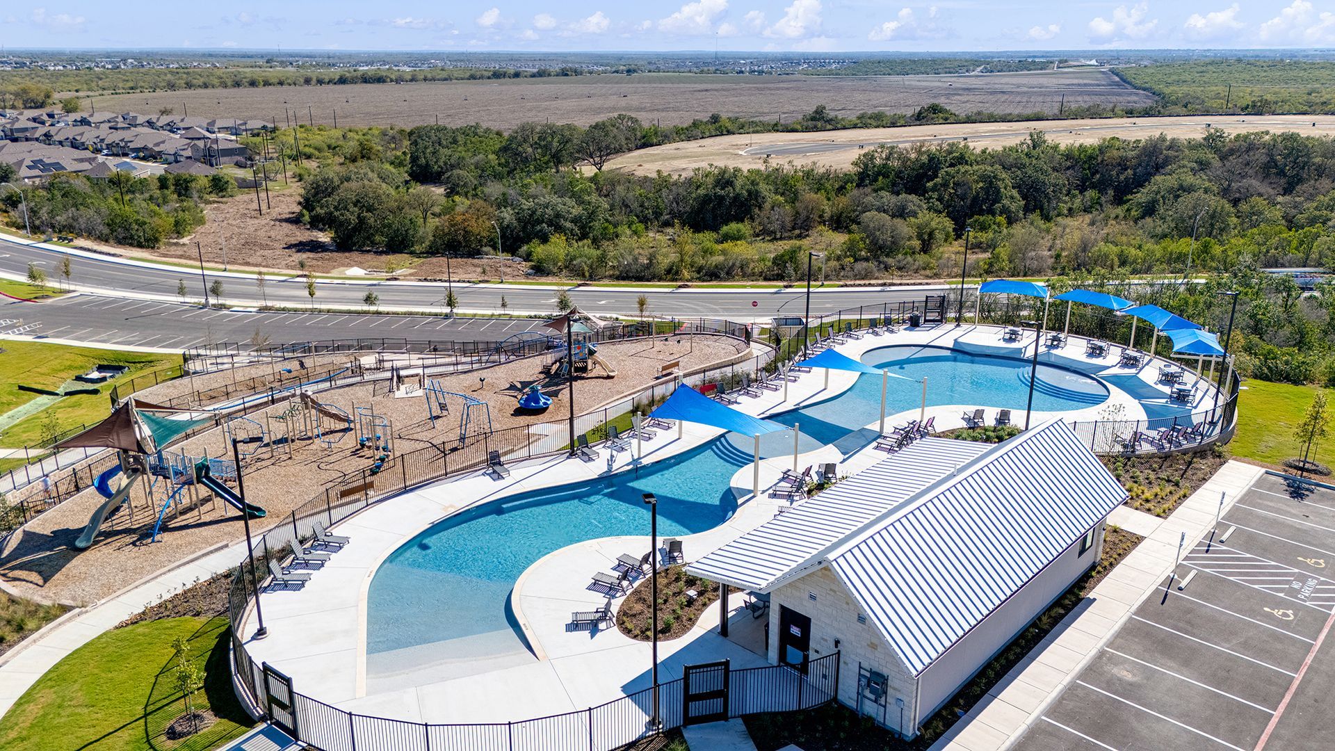 An aerial view of a swimming pool complex with a sandy playground, lounge chairs, and a small building in a sunny area.