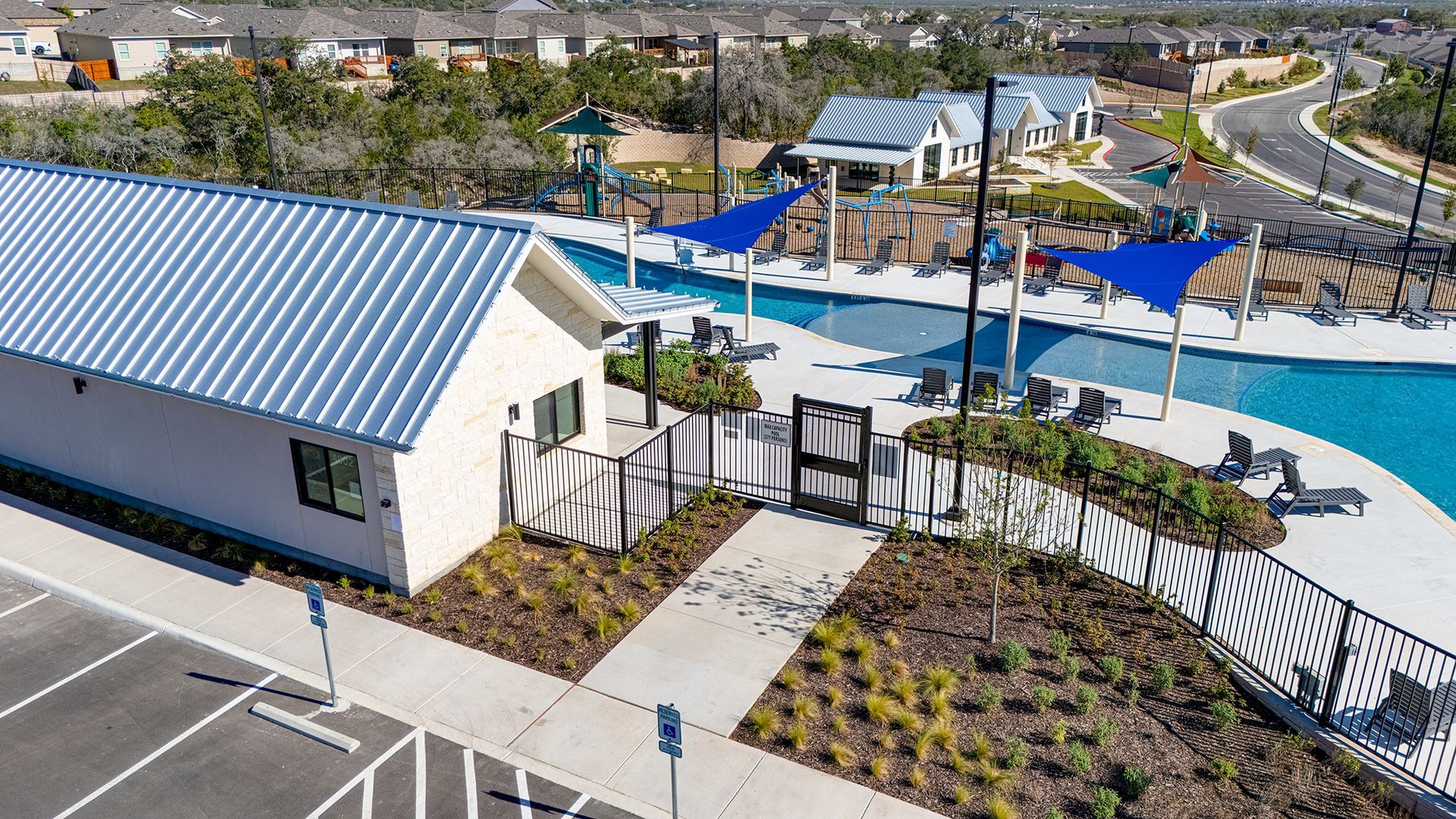 Aerial view of a community pool facility with a building, swimming lanes, blue shade sails, and an adjacent parking lot.