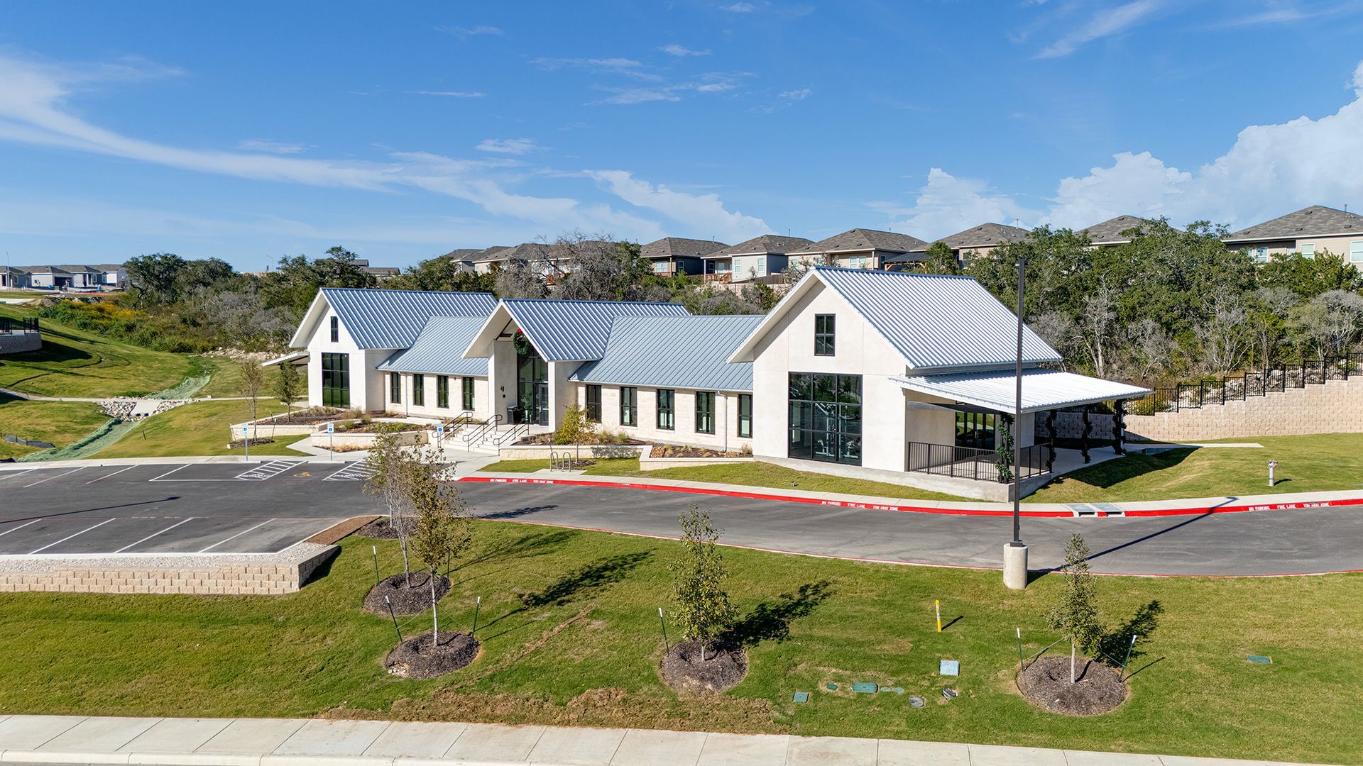 A modern, one-story white building with a gray metal roof, situated near a parking lot with a manicured lawn and trees.